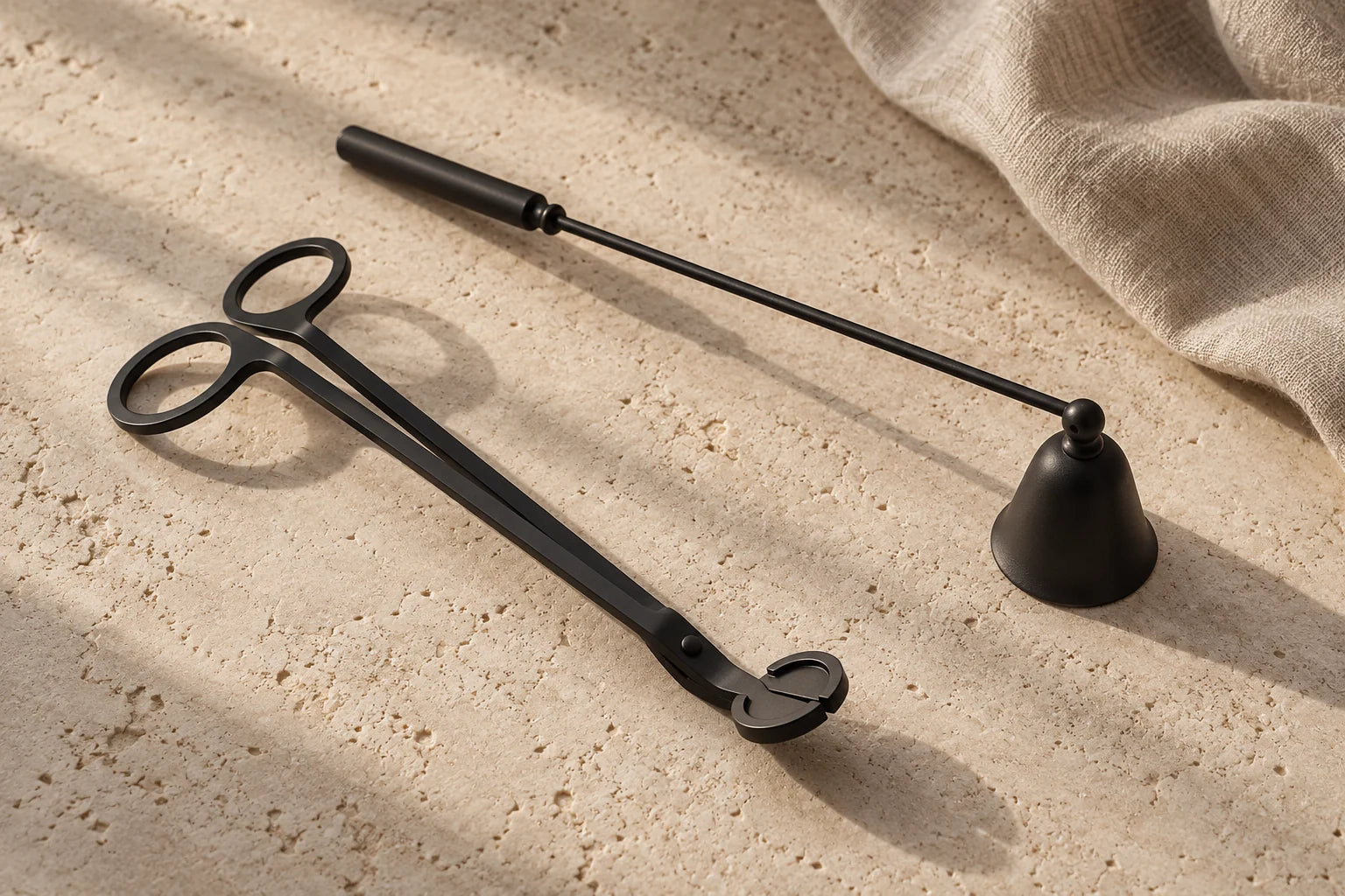 Black wick trimmer and candle snuffer resting on warm limestone beside linen